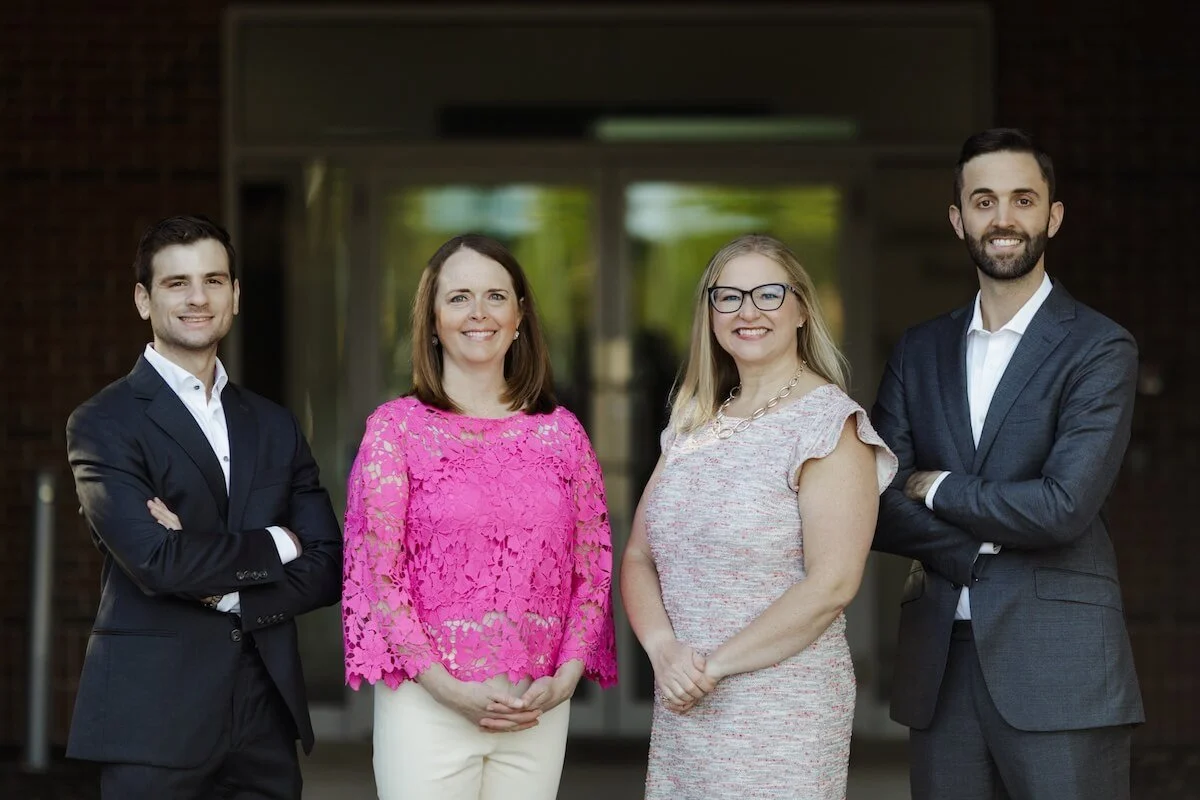 Four professionally dressed people standing outdoors in front of a building, smiling at the camera.