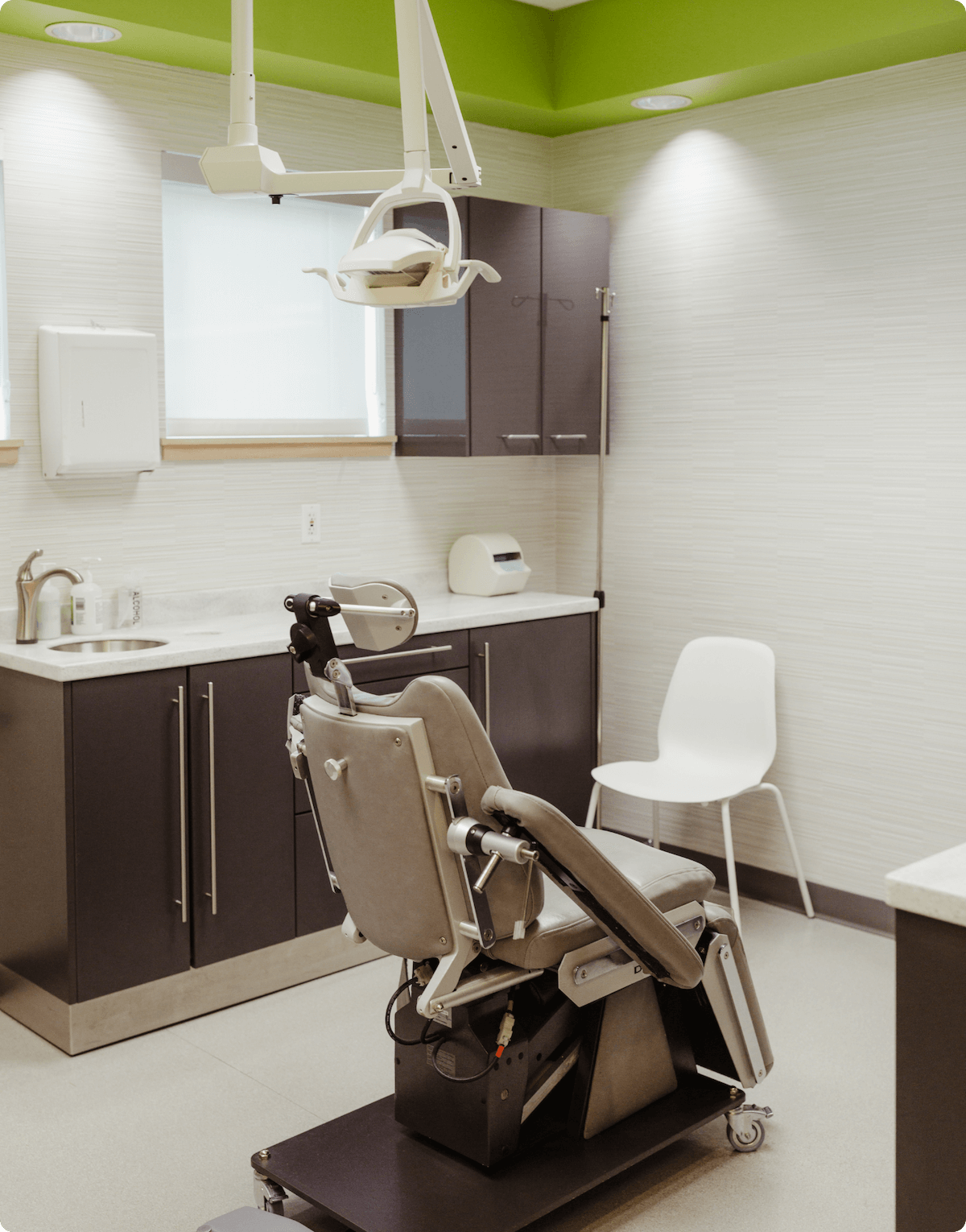 An empty medical examination room with a gynecological examination chair, medical equipment, cabinets, and a white chair in the background.