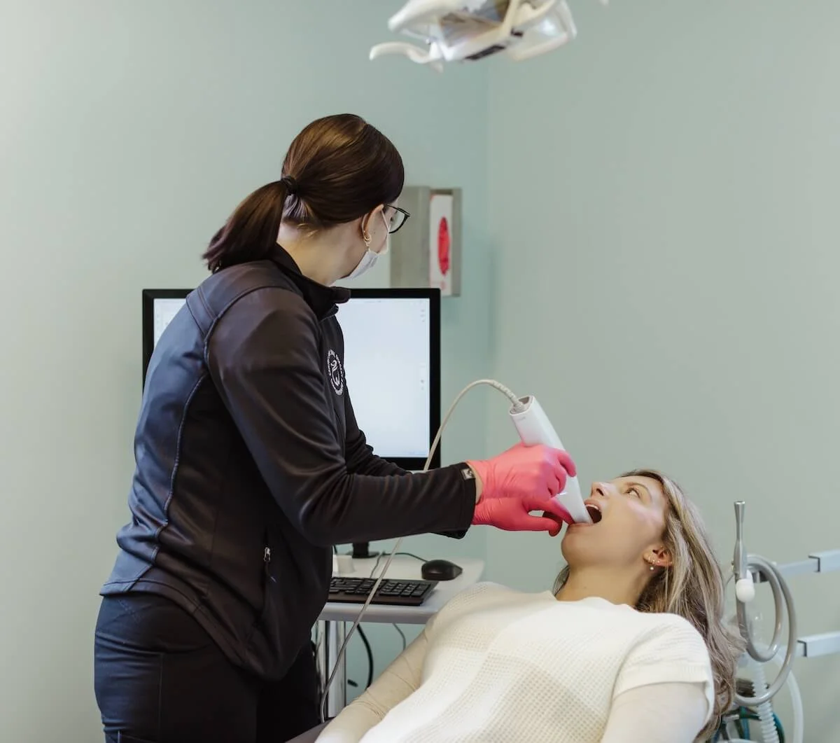A woman in a black jacket and pink gloves performs an oral exam with a scanning device on a patient lying in a dental chair in a clinical setting.
