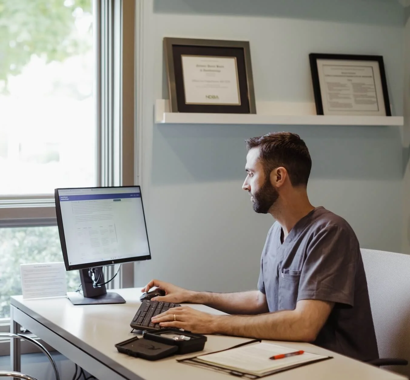 A man in medical scrubs working at a computer in an office, with framed certificates on the wall behind him.