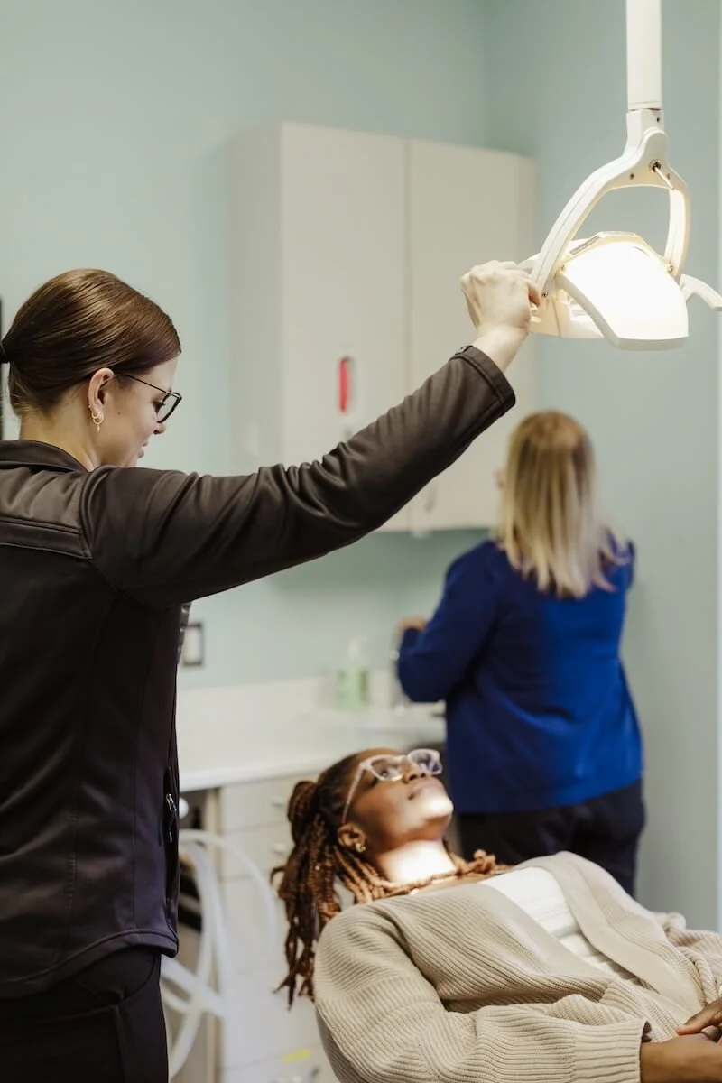 A woman lying back in a dental chair with her eyes closed, a healthcare professional adjusting an overhead light in a dental office.