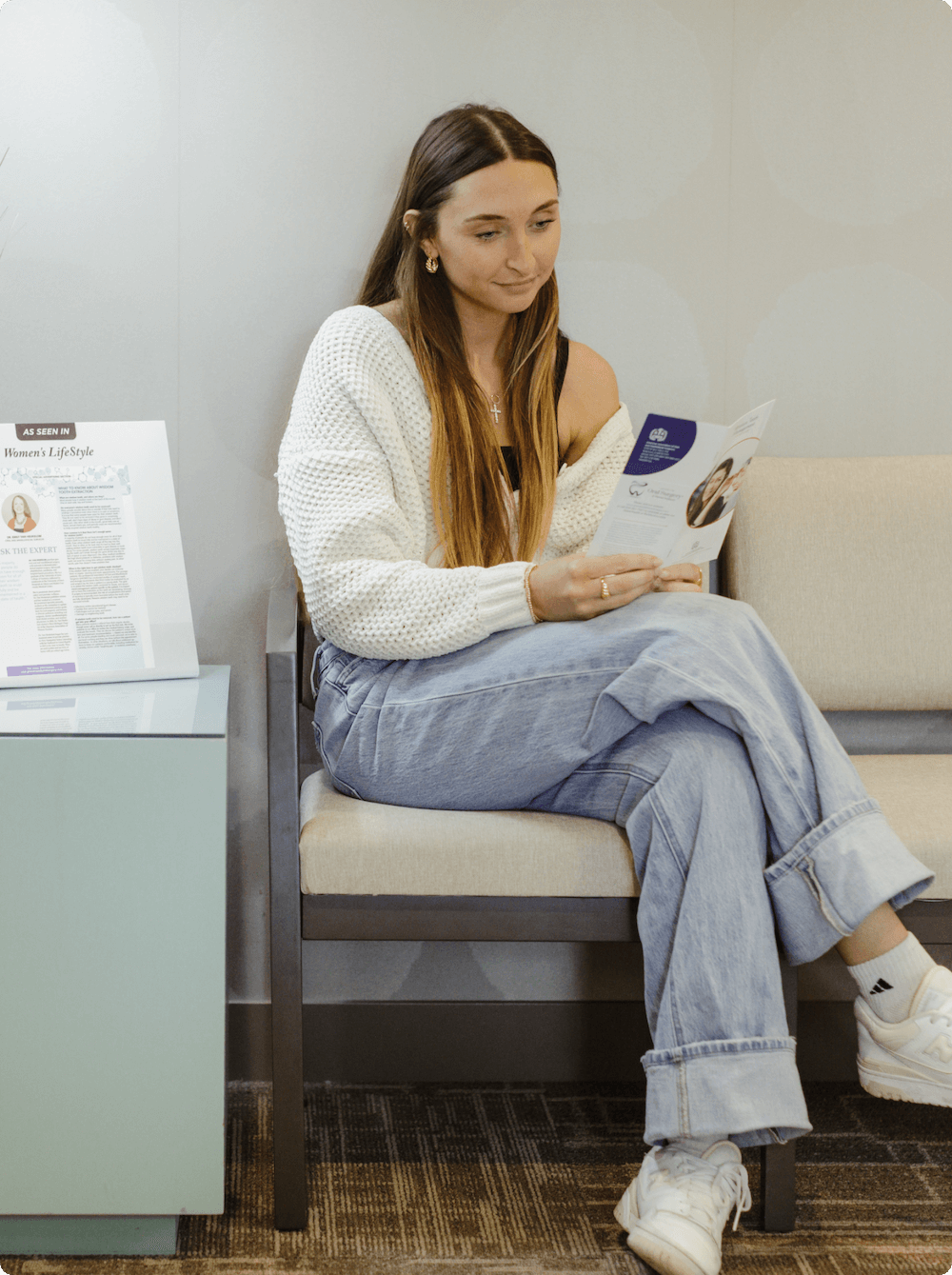 A young woman with long brown hair, wearing a white sweater, black tank top, light blue jeans, and white sneakers, sitting on a waiting room bench and reading a pamphlet.