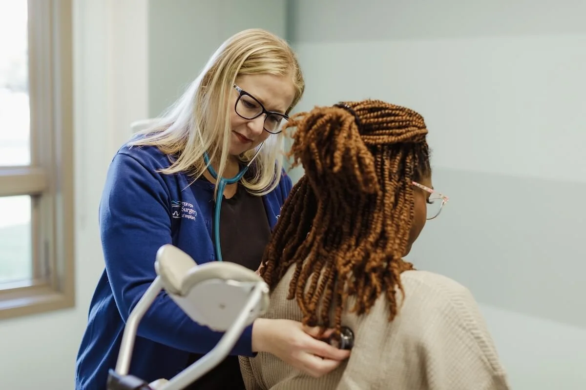 A healthcare professional with blonde hair, wearing glasses and a blue jacket, examining a patient's back with a stethoscope in a clinic room.