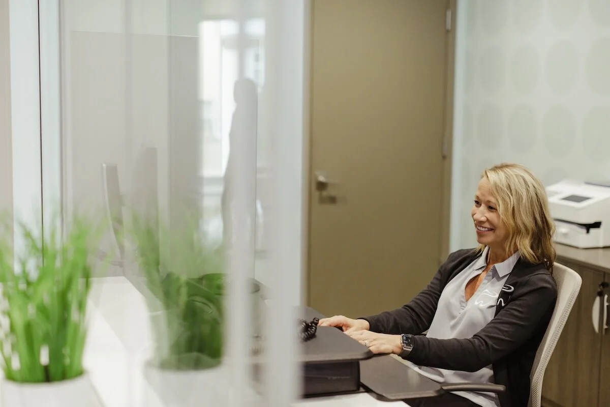 A woman with blonde hair working at a desk in an office, smiling, with a computer monitor and a potted plant in the foreground.