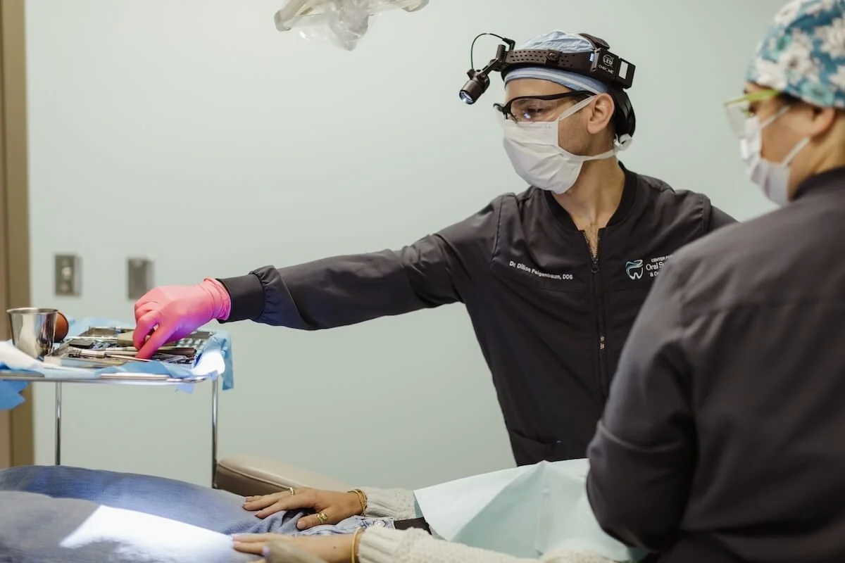 A healthcare professional in surgical scrubs, wearing a mask, glasses, and a headlamp, is performing a dental procedure on a patient in a clinical setting.