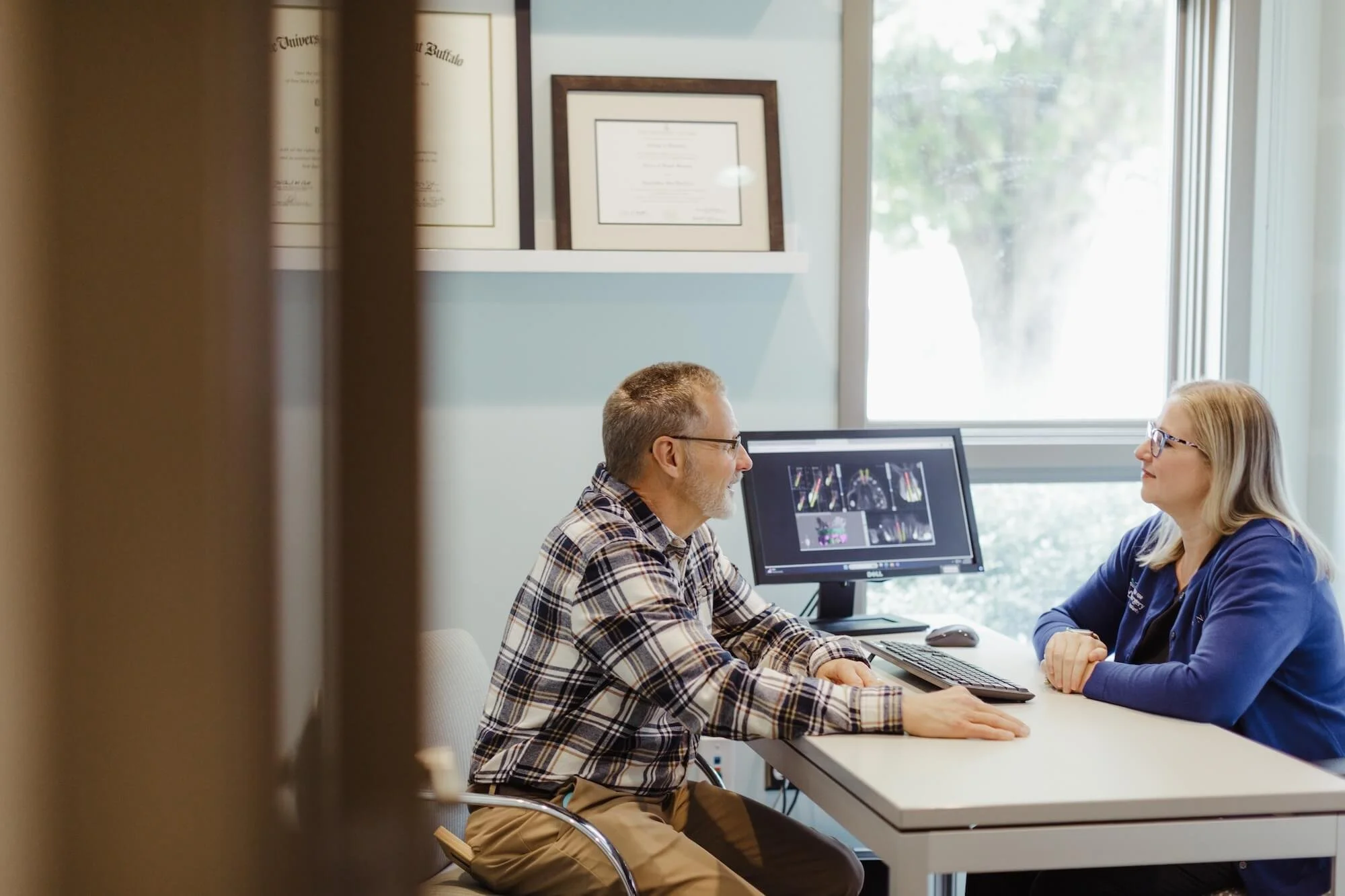 A doctor and a patient sitting across from each other at a desk in a medical office, discussing medical images on a computer monitor.