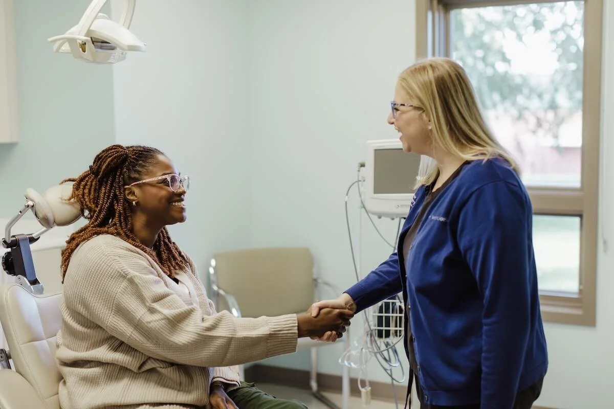 A healthcare professional shaking hands with a female patient in a medical examination room, both smiling.