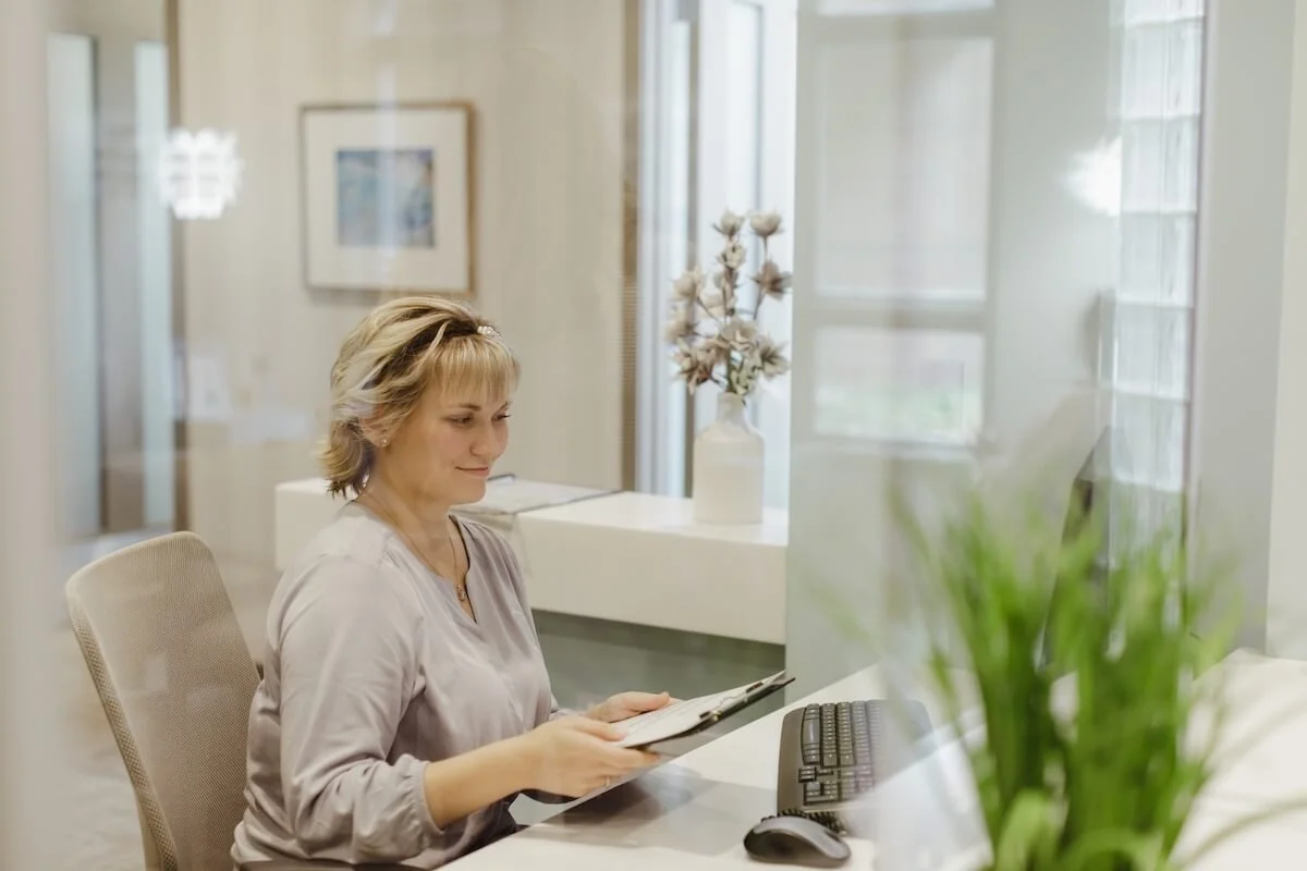 A woman sitting at a desk, looking at a tablet, in a modern, well-lit office or waiting area with a window and decorative objects in the background.