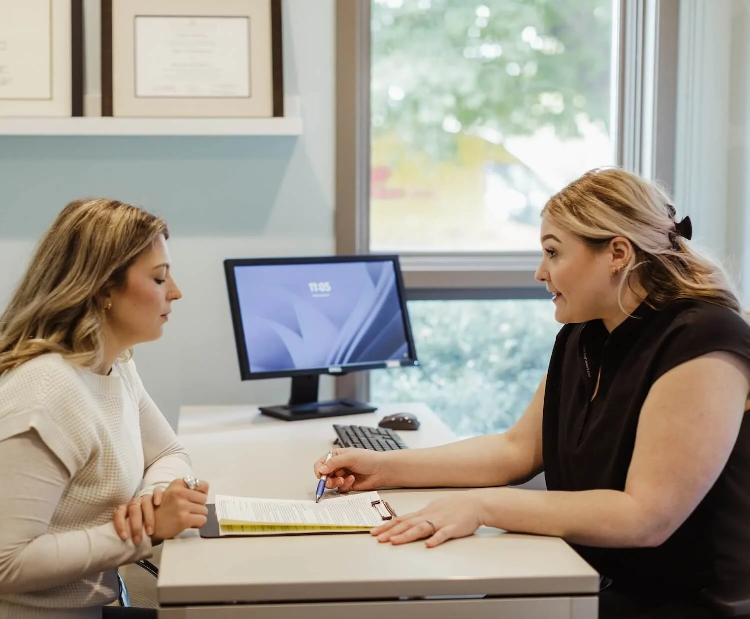 Two women sit across from each other at an office desk, engaged in a discussion. One woman is taking notes with a pen on a notepad, while the other listens attentively. A computer monitor is on the desk, and framed certificates or diplomas are hanging on the wall behind them. Large windows behind them let in natural light.