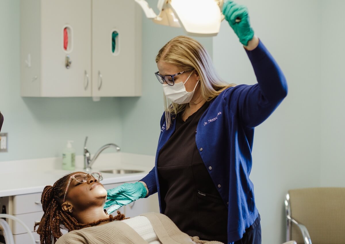 A female doctor or dentist wearing a mask, gloves, and scrubs examining a patient lying on a medical chair in a clinical setting.