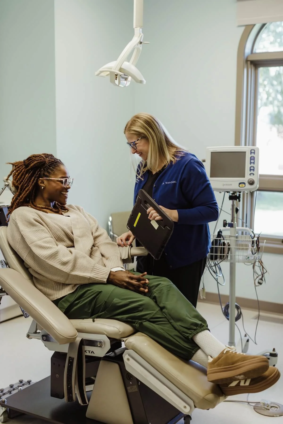 A woman sitting in a hospital bed talking to a nurse in a medical room with monitors and medical equipment.