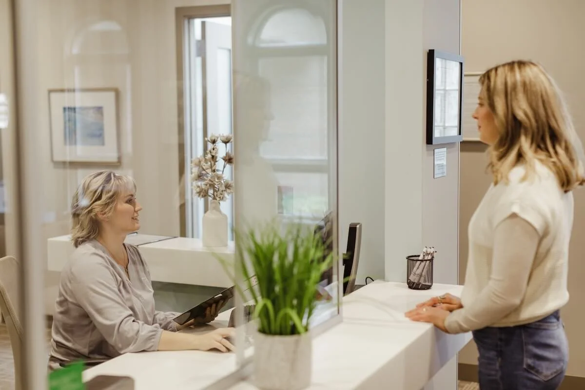 A woman checking in at a reception desk with a receptionist seated behind a glass partition, holding a tablet, in a modern office or clinic setting.