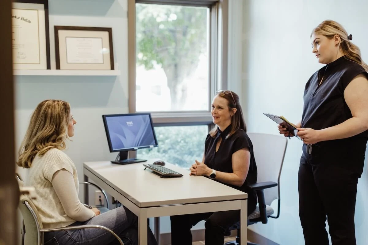 A woman sitting across a desk from a talking therapist or counselor during a therapy session in an office with framed certificates on the wall near a window.