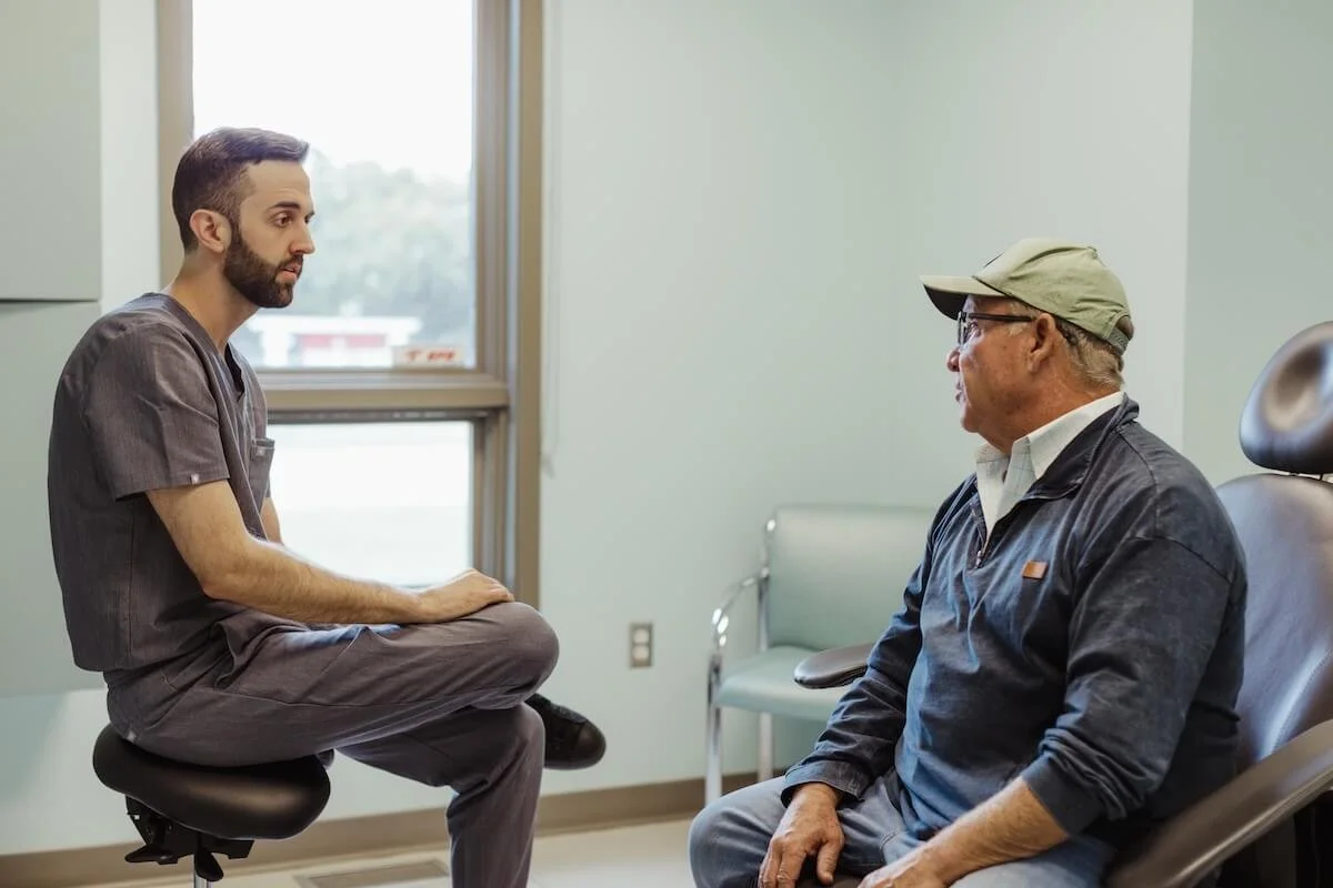 A young man in medical scrubs sits cross-legged on an examination stool, facing an older man wearing glasses, a cap, and a dark jacket, who is seated in a medical examination chair.