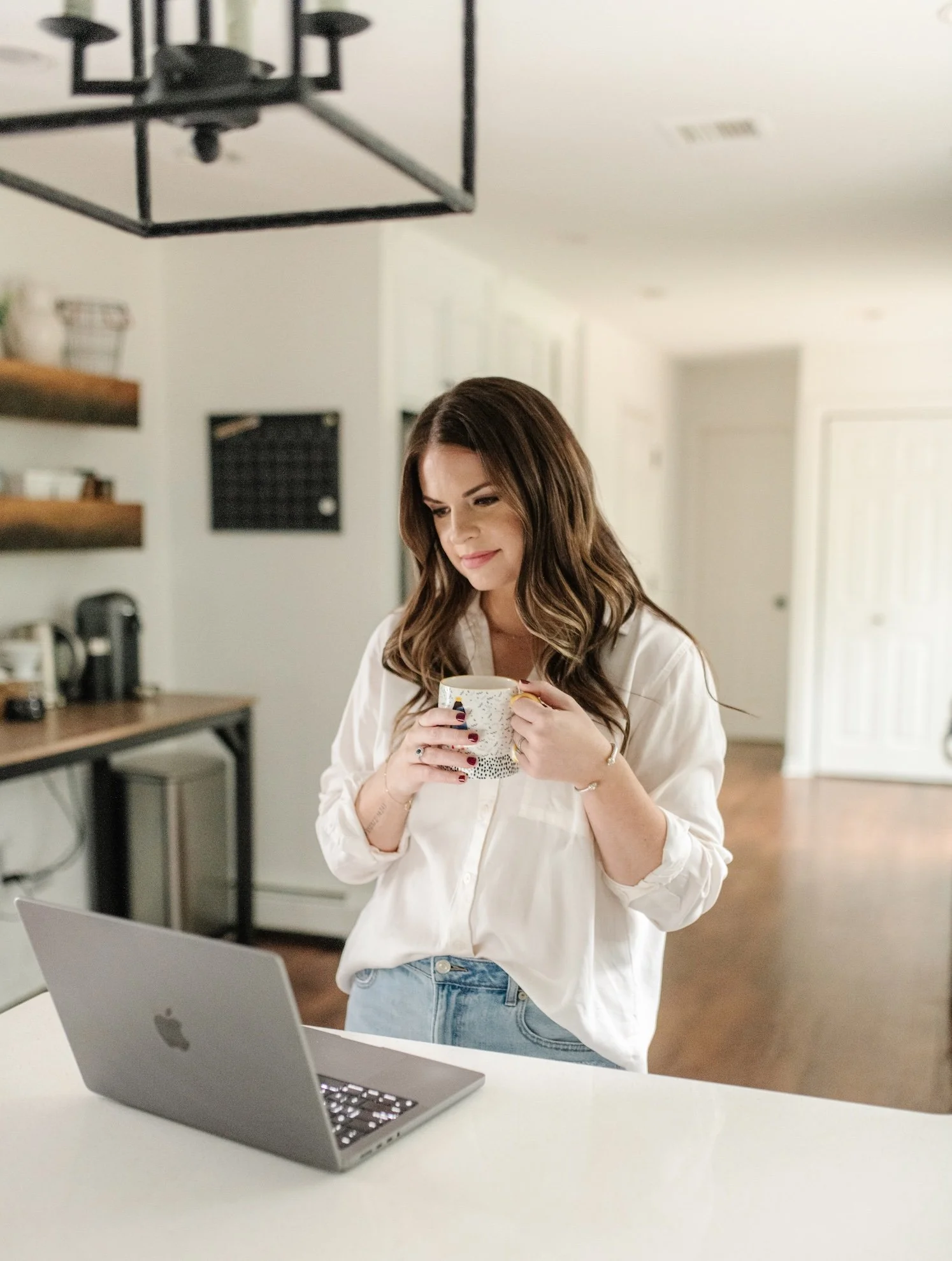 A woman with long brown hair, wearing a white blouse and jeans, is standing at a table holding a mug and looking down. There is a silver laptop in front of her on the table. The background shows a kitchen with shelves, a coffee machine, and a laptop.