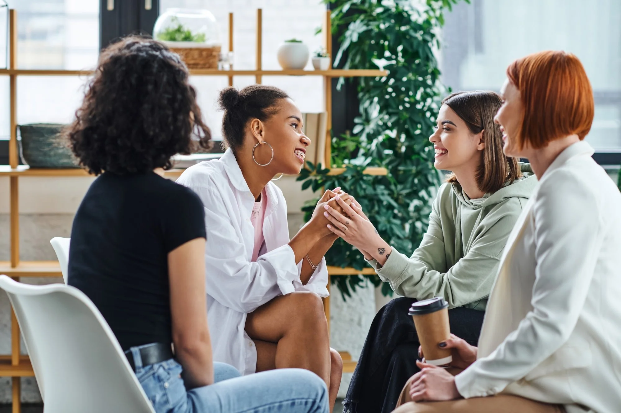 Four women sitting in a circle engaged in conversation in a bright room with large windows and plants.