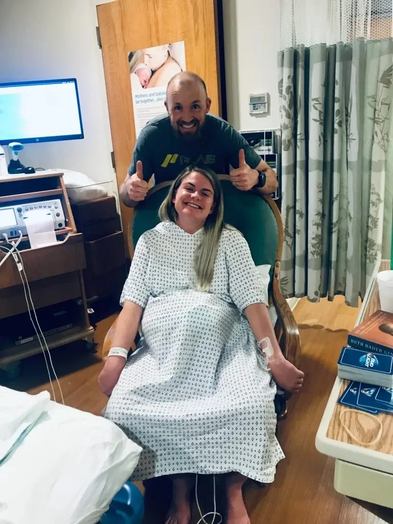 A woman in a hospital gown sitting in a hospital bed, smiling, with IV lines in her arms. A man standing behind her in a hospital room giving two thumbs up and smiling.