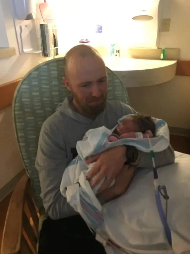 A man sitting in a hospital room holding a newborn baby wrapped in a blanket.