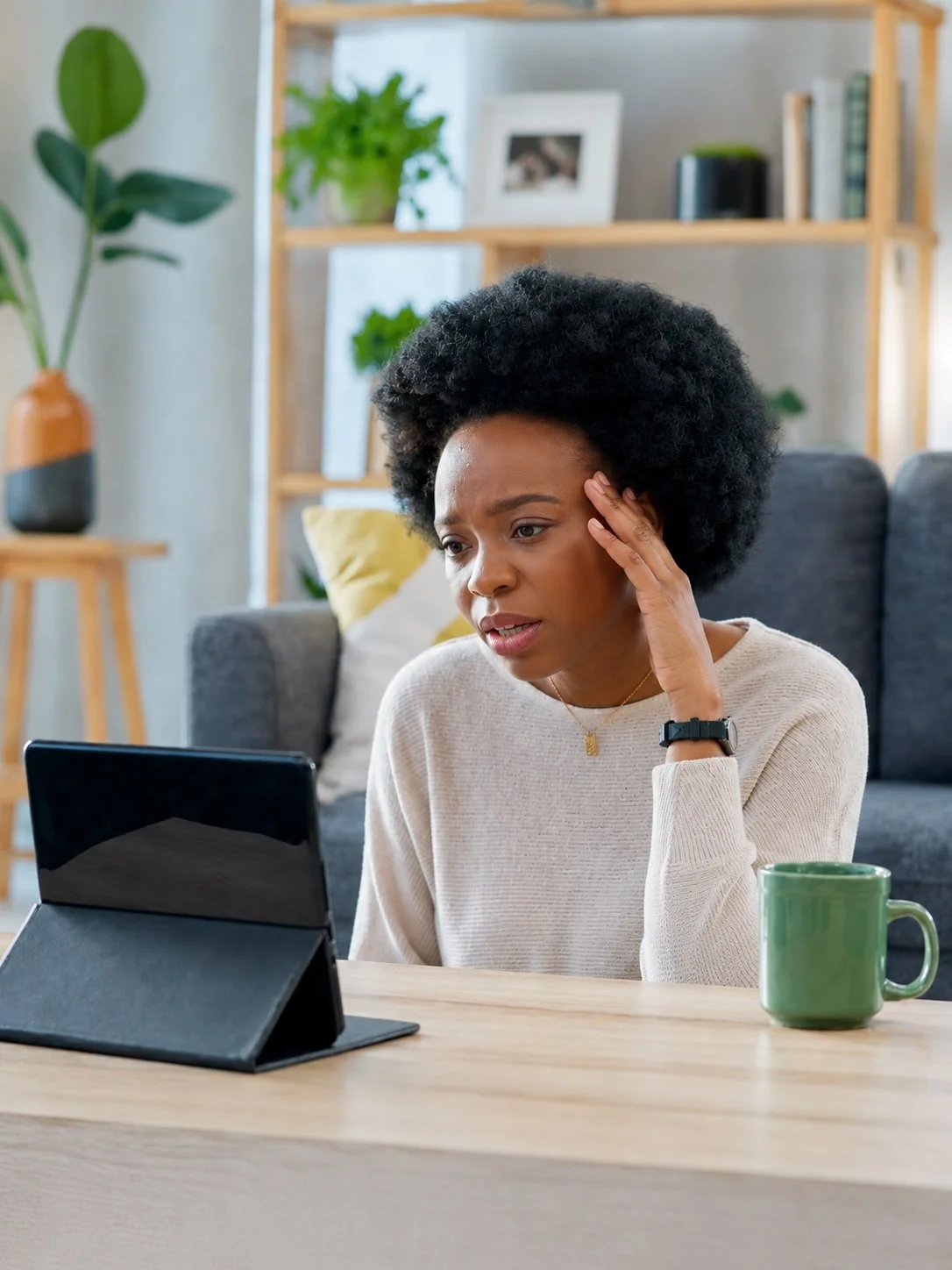 A woman sitting at a wooden table, looking concerned at a tablet in front of her, with a green mug beside it, in a cozy living room with a gray couch, bookshelf, and houseplants in the background.