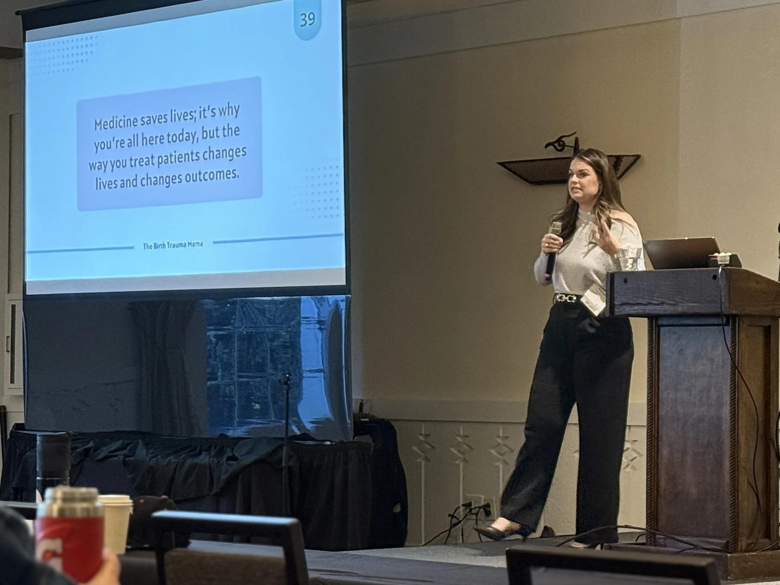 Kayleigh Summers giving a presentation in front of a projection screen, with coffee cups on the table in the foreground. The slide on the screen discusses the importance of medicine in saving lives and changing patient outcomes.