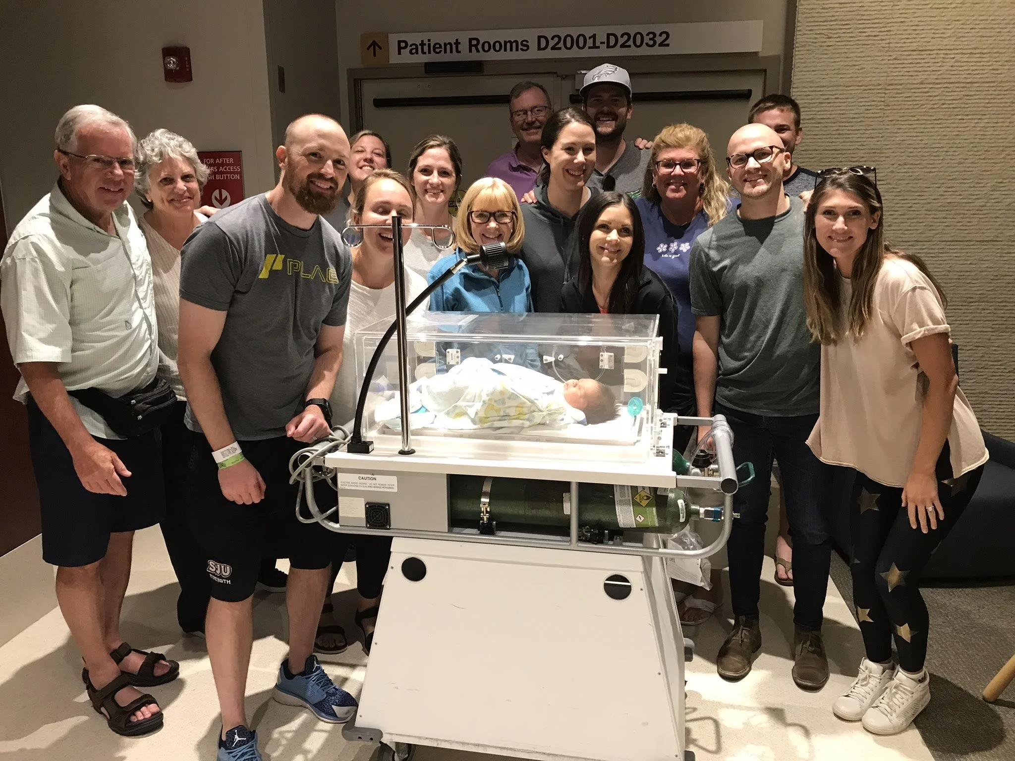 A group of people including men and women smiling around a hospital baby incubator with a newborn inside, in a hospital corridor. There is a sign overhead indicating patient rooms and a red safety button on the wall.