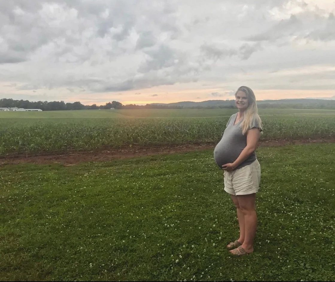 Pregnant woman standing outdoors on grassy field during sunset, with a green farmland background and cloudy sky.