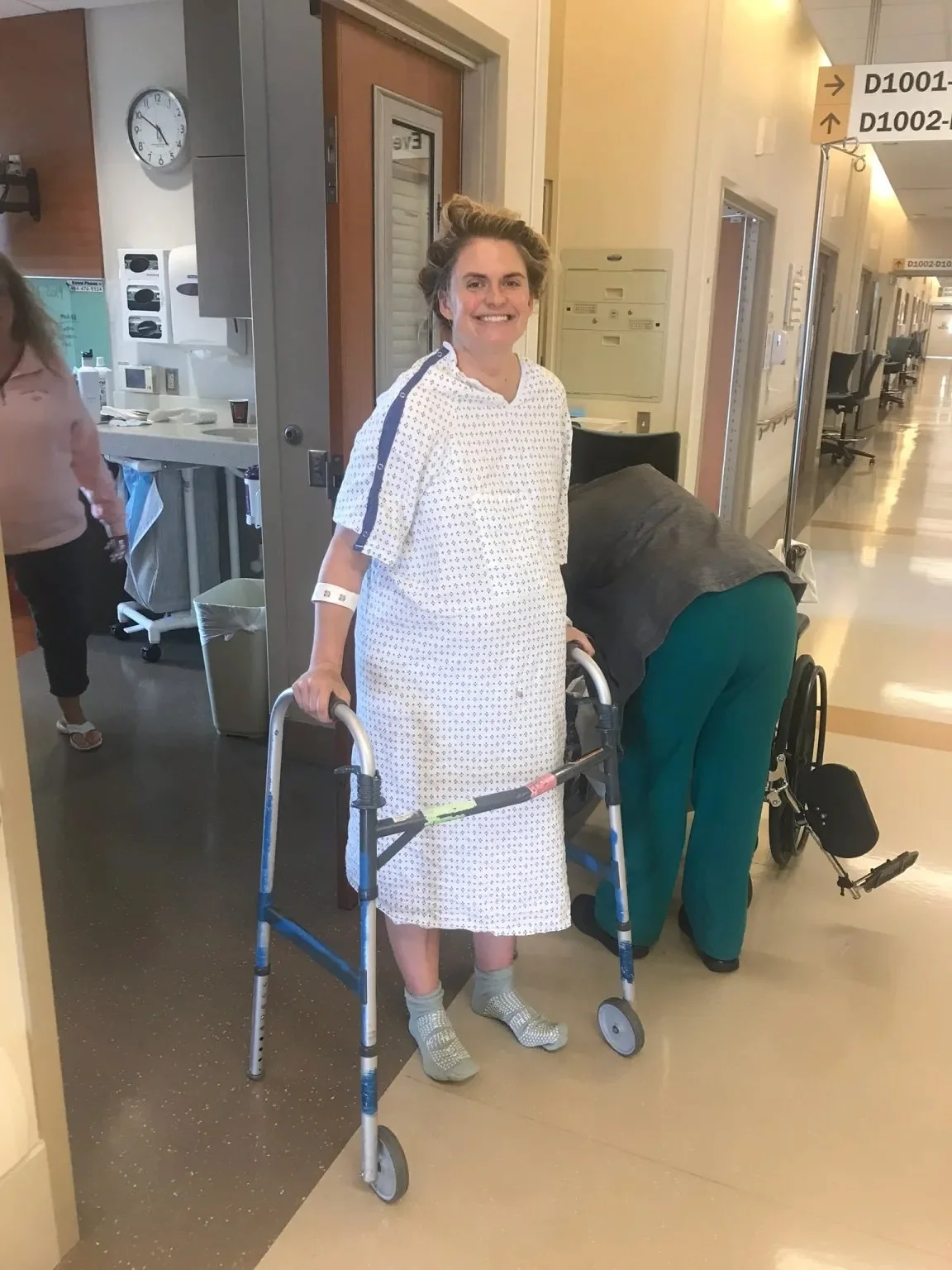 A smiling woman in a hospital gown stands with a walker in a hospital corridor.
