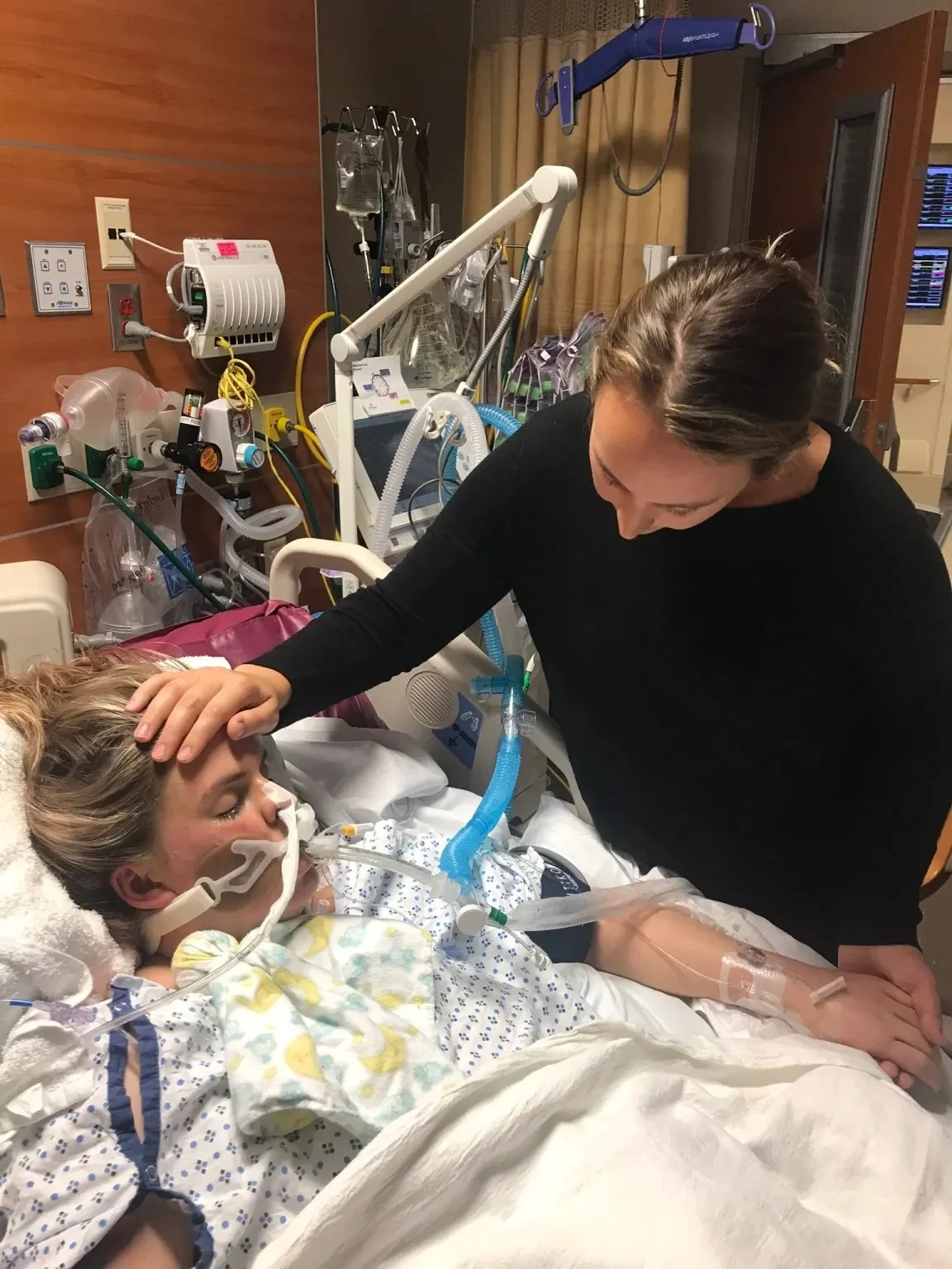 A woman in a hospital bed with a breathing mask and medical equipment, receiving comfort from a visitor who is touching her forehead.