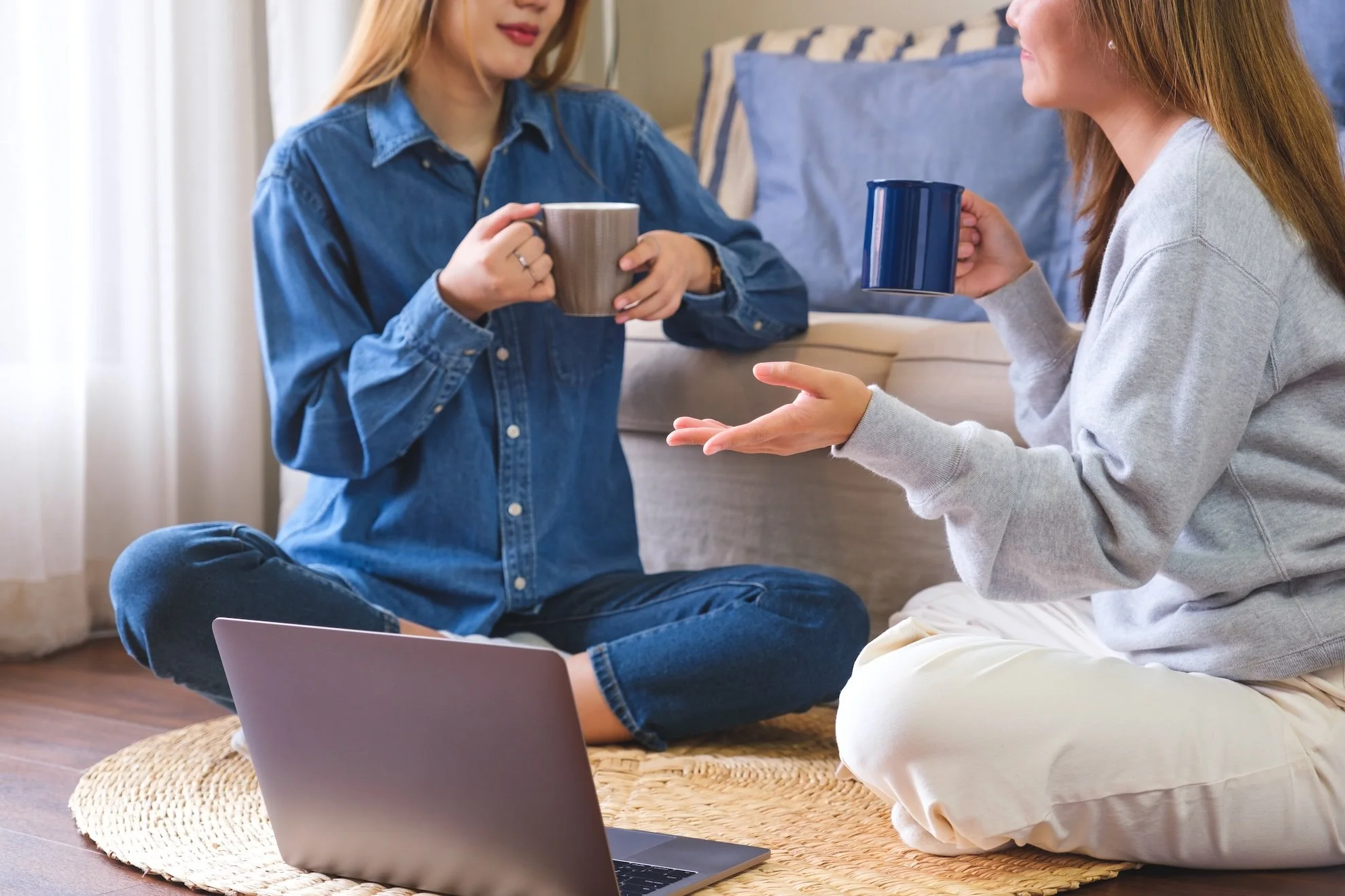 Two women sitting on a woven rug in a cozy room, engaging in a conversation while holding coffee mugs. One woman has long red hair, and the other has long blonde hair. A laptop is open in front of them.