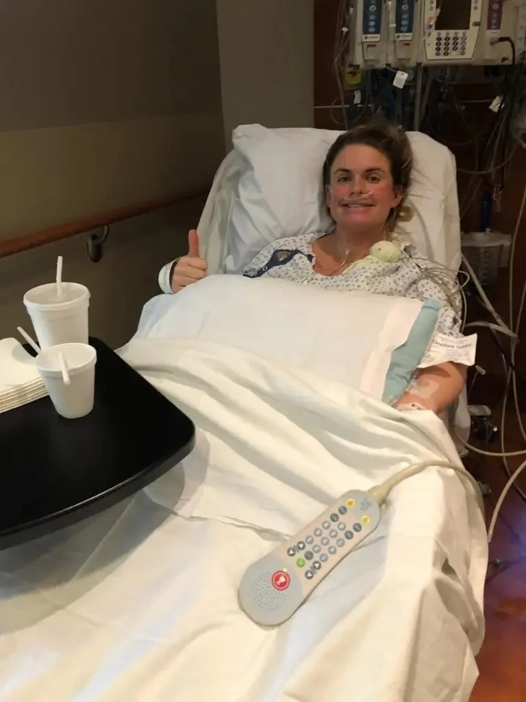 Woman in hospital bed smiling and giving a thumbs-up, with medical equipment and drinks nearby.