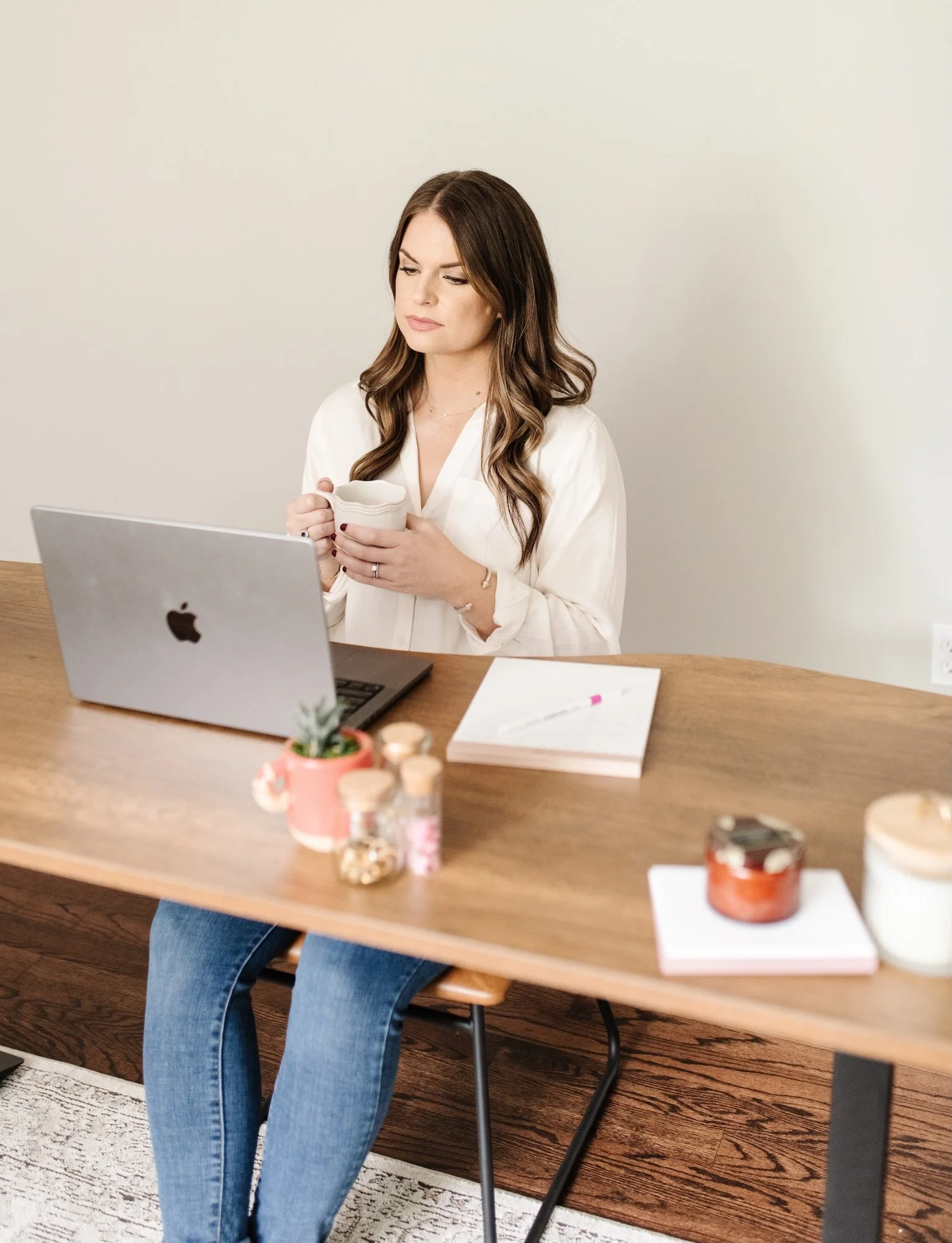 A woman with long brown hair sitting at a wooden table, looking at her phone, with an open laptop in front of her and various small jars and a notebook on the table.