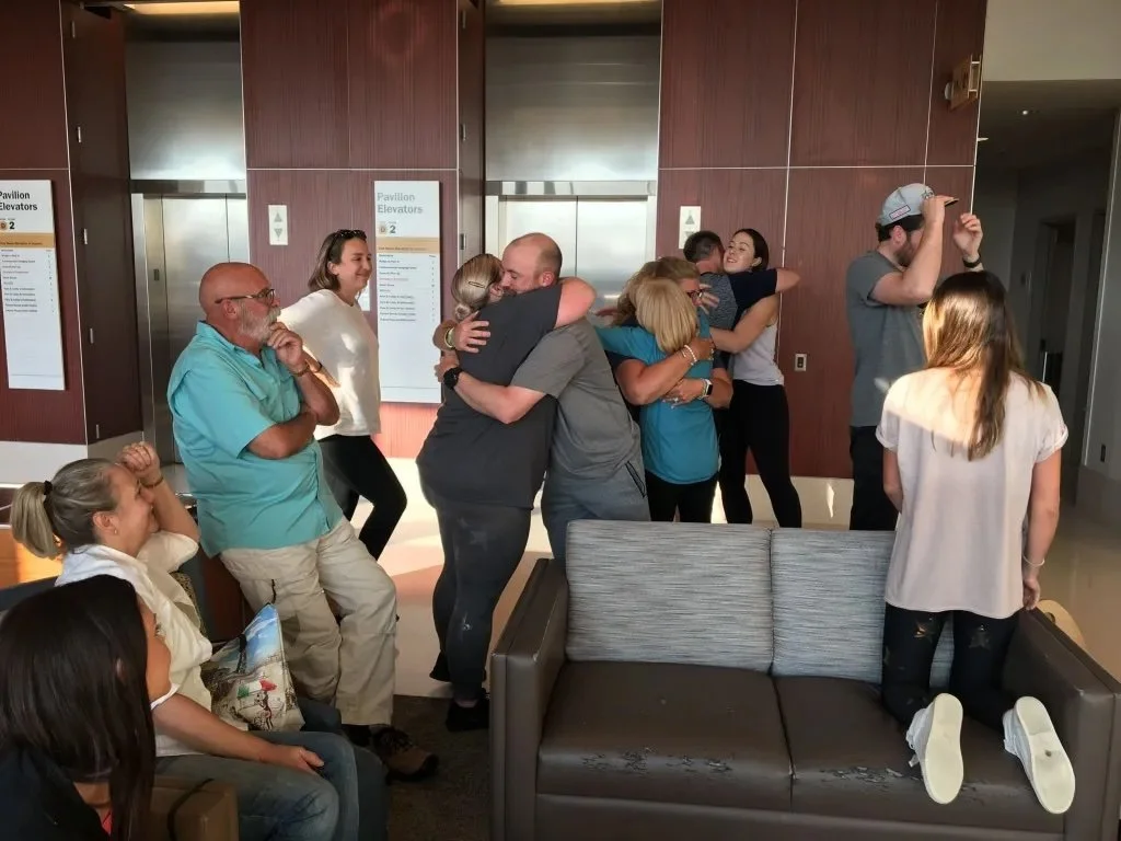 Group of people hugging and socializing in a lobby with elevators, some sitting and others standing.