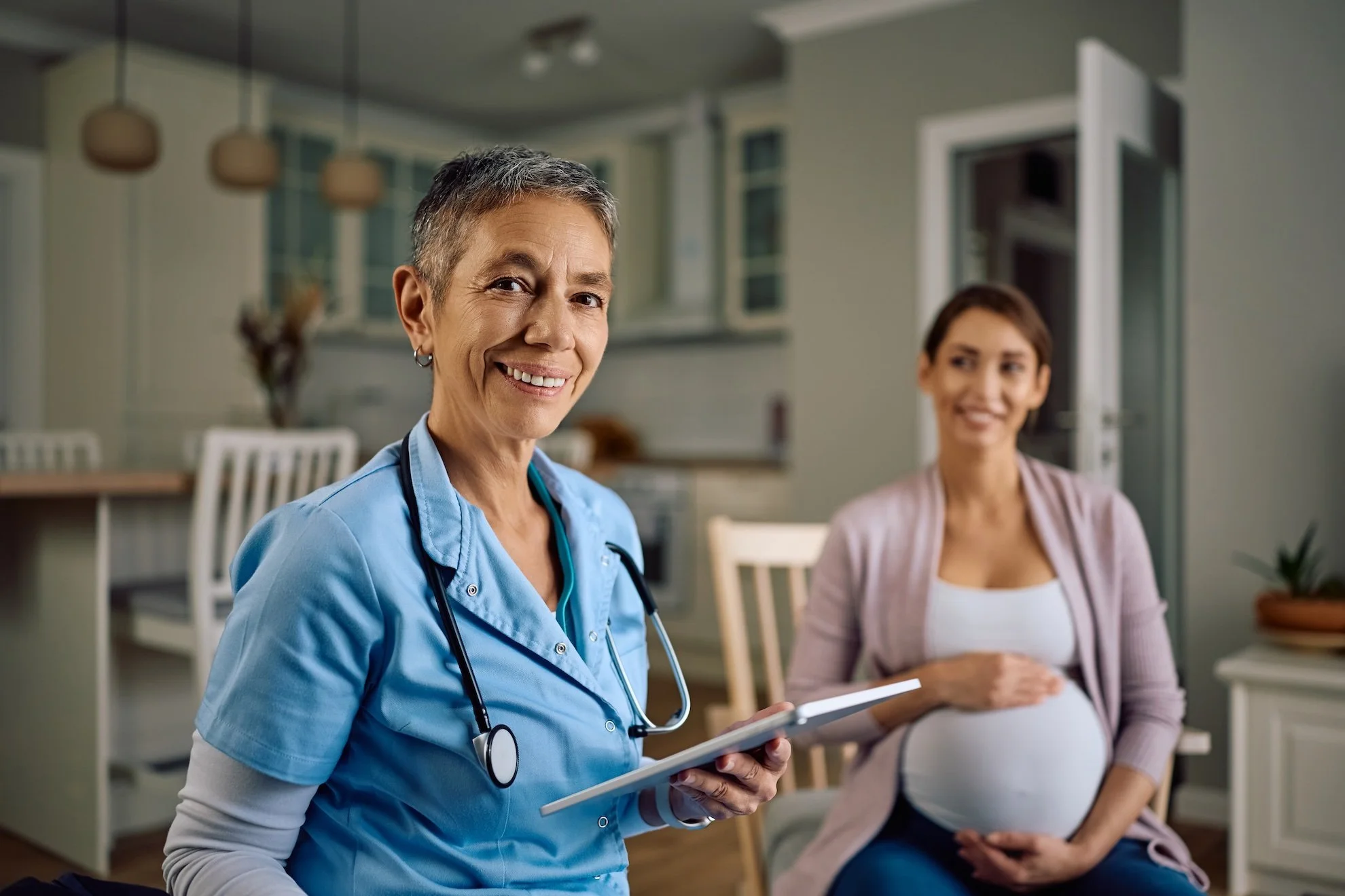 A female healthcare professional in scrubs holding a tablet, smiling at the camera, with a pregnant woman sitting behind her and to the right, inside a modern home or clinic.