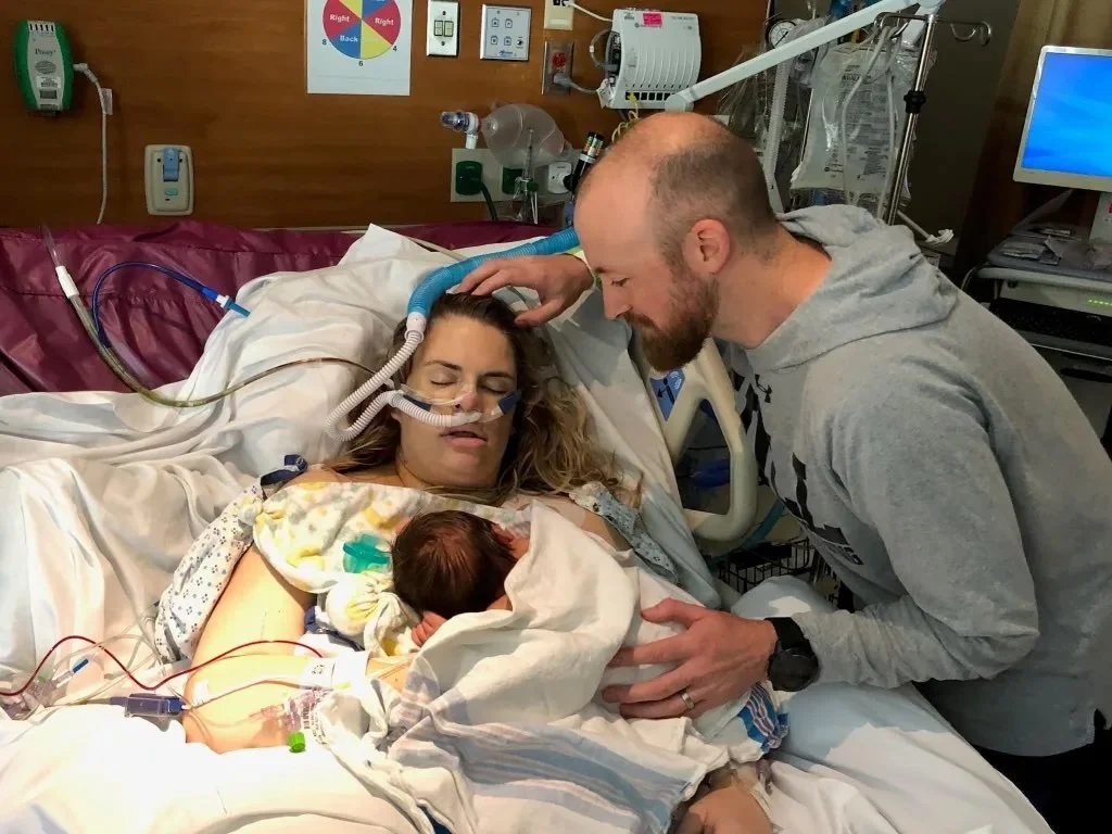 A woman in a hospital bed holding her newborn baby, with a man (likely her partner) leaning over her, both looking at the baby. The woman is connected to medical tubes and monitors, indicating she is in a medical setting, probably a hospital.