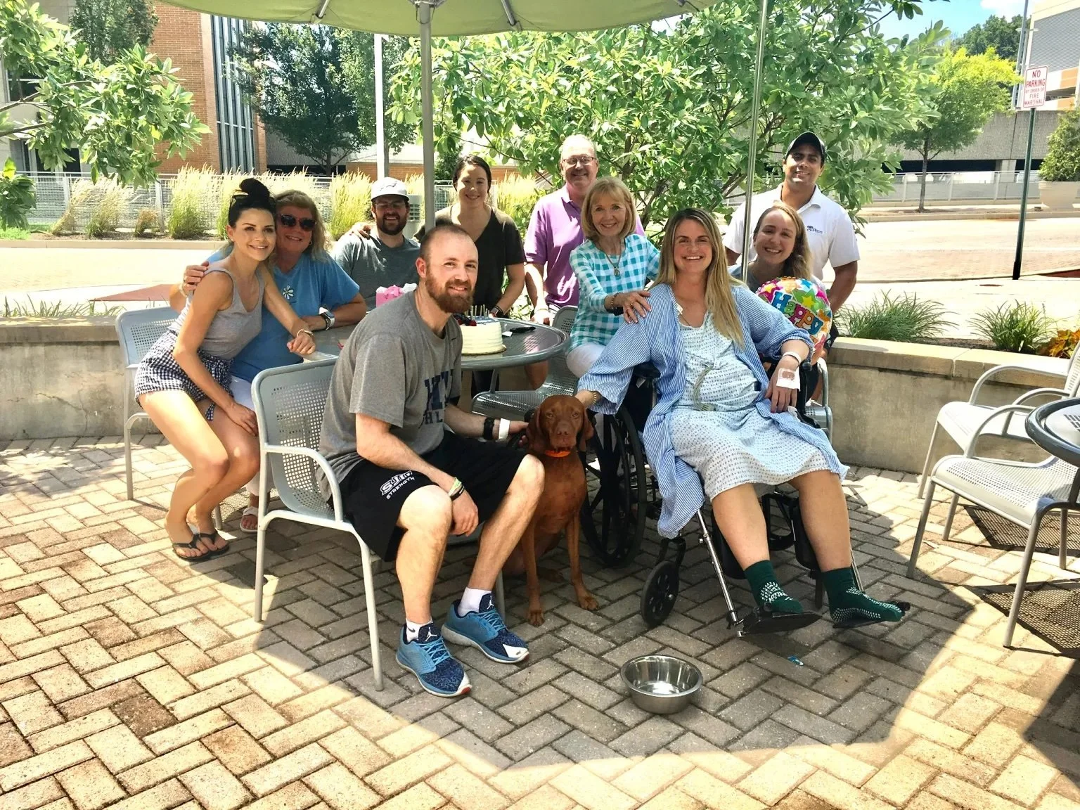A group of people gathered outdoors around a table under a green umbrella, celebrating a birthday. There is a woman in a hospital gown sitting in a wheelchair, a man beside her, and a dog sitting between them. The group is smiling and posing for the photo in a sunny setting with trees, brick pavement, and a building in the background.