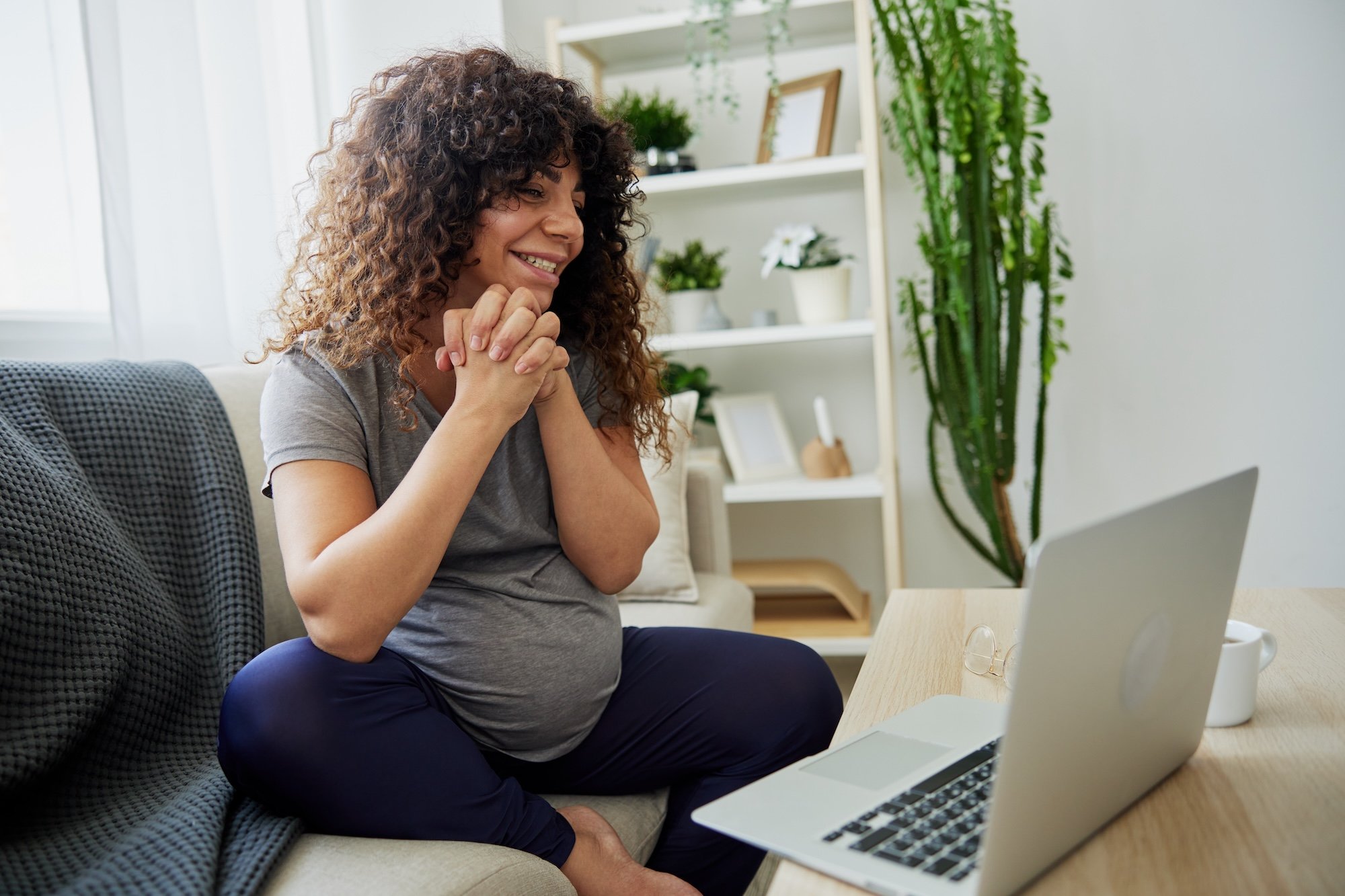 A woman with curly brown hair sitting on a sofa in front of a laptop, smiling, with her hands clasped under her chin. There are plants and shelves in the background, with sunlight coming through a window.