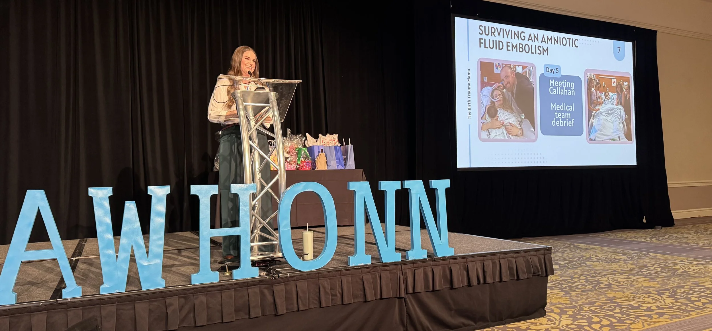 A woman standing at a podium on a stage with large blue letters spelling 'AWHHON' in front of her, presenting at a conference. Behind her, a large screen shows a slide titled 'Surviving an Amniotic Fluid Embolism' featuring two images and a timeline with the step 'Meeting Callahan Medical Team debrief' on Day 5.