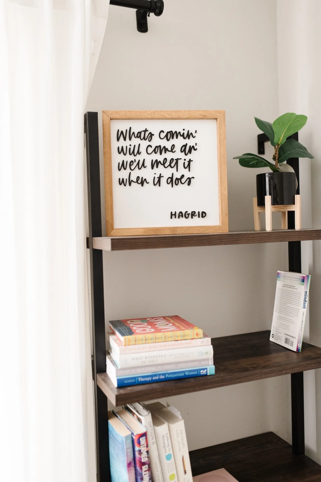 A bookshelf with five levels contains books, a small potted plant, a framed quote, and a pamphlet.