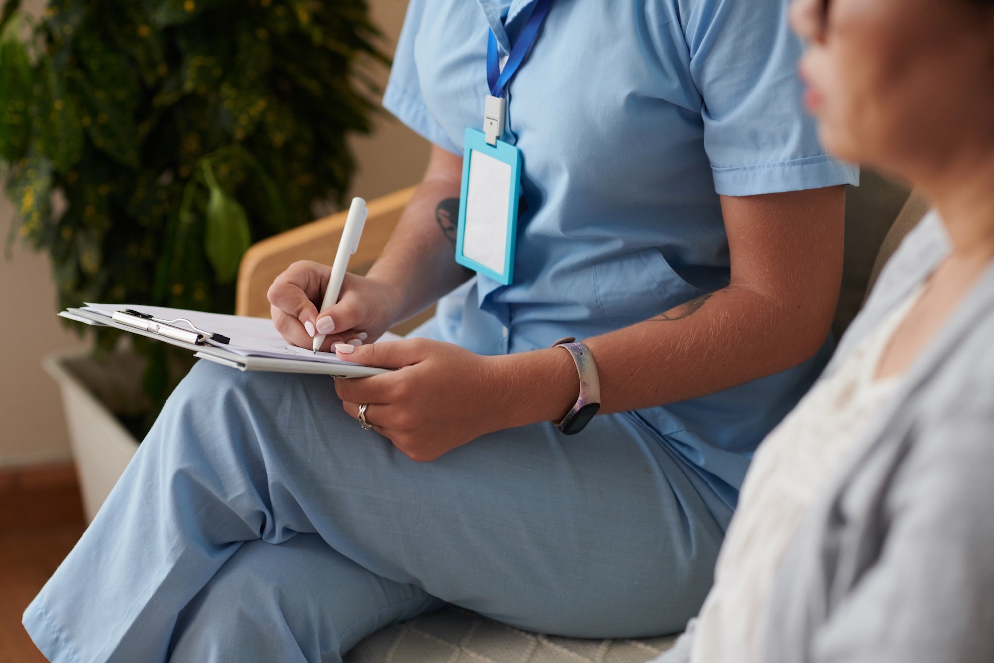 A healthcare professional sitting with a patient, taking notes on a clipboard, wearing scrubs and a medical ID badge.