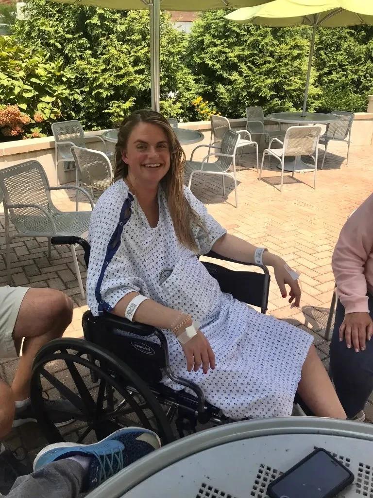 Woman in hospital gown sitting outside in a wheelchair on a patio, smiling, with greenery and patio tables in the background.