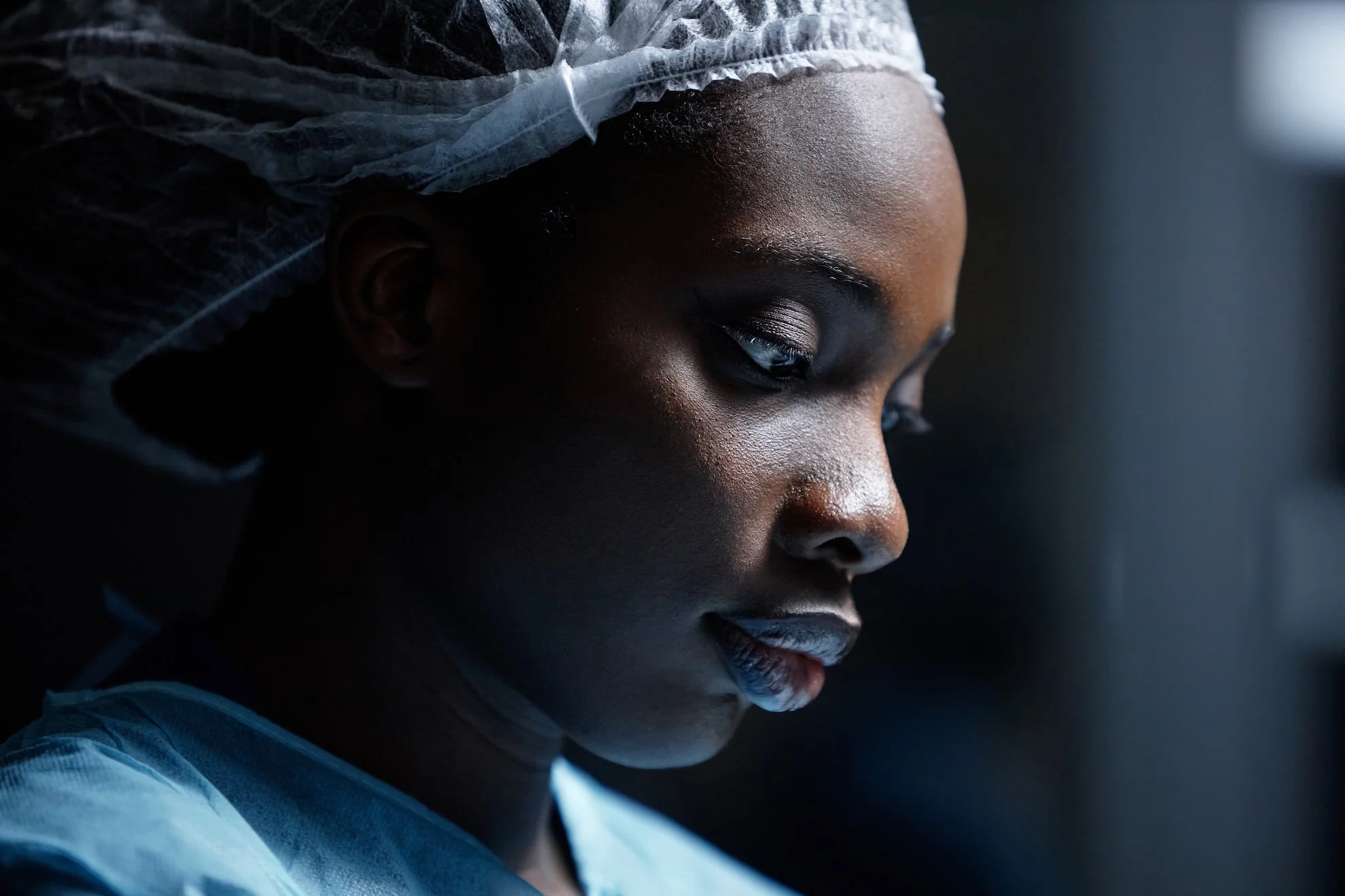 Close-up of a woman with dark skin and closed eyes, wearing a white medical gown and a disposable hair cover, in a professional medical setting.