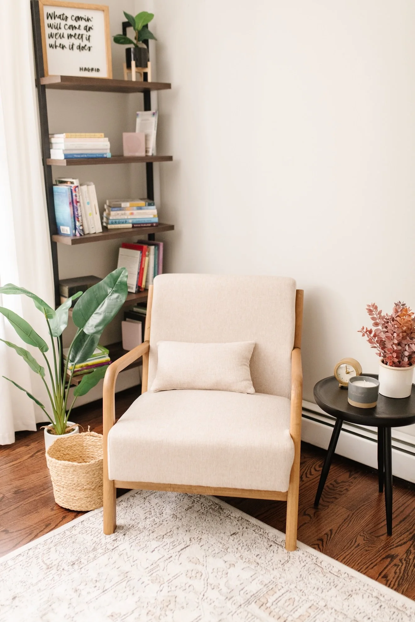 A cozy living room corner featuring a beige armchair with a matching pillow, a black side table with candles and a plant, a tall shelving unit with books and decor, indoor plants, and a white rug with a subtle pattern. There is a framed quote on the top shelf and a basket next to the chair.
