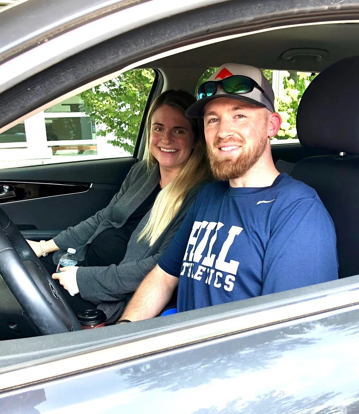 A man and woman sitting inside a car, smiling for a photo. The woman is holding a water bottle, and the man is wearing sunglasses on his cap. Outside the car window, trees and a building are visible.