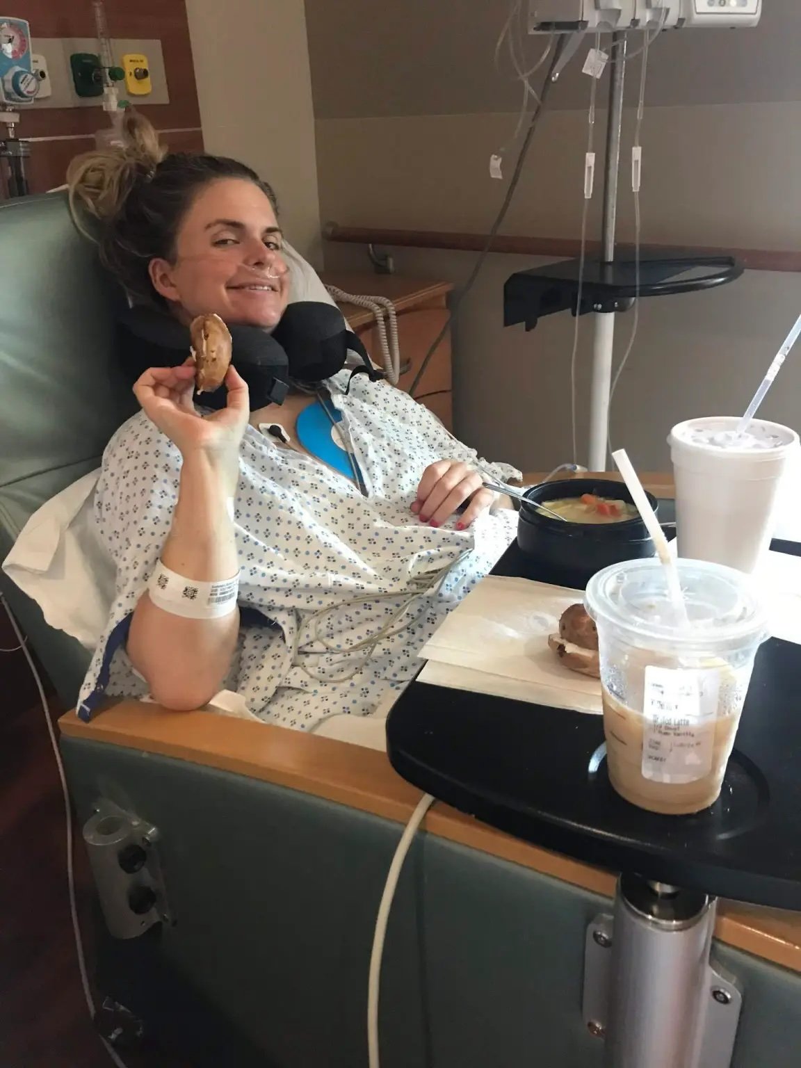 Hospital patient in a hospital bed holding a cookie and smiling, with drinks and food on a tray in front.