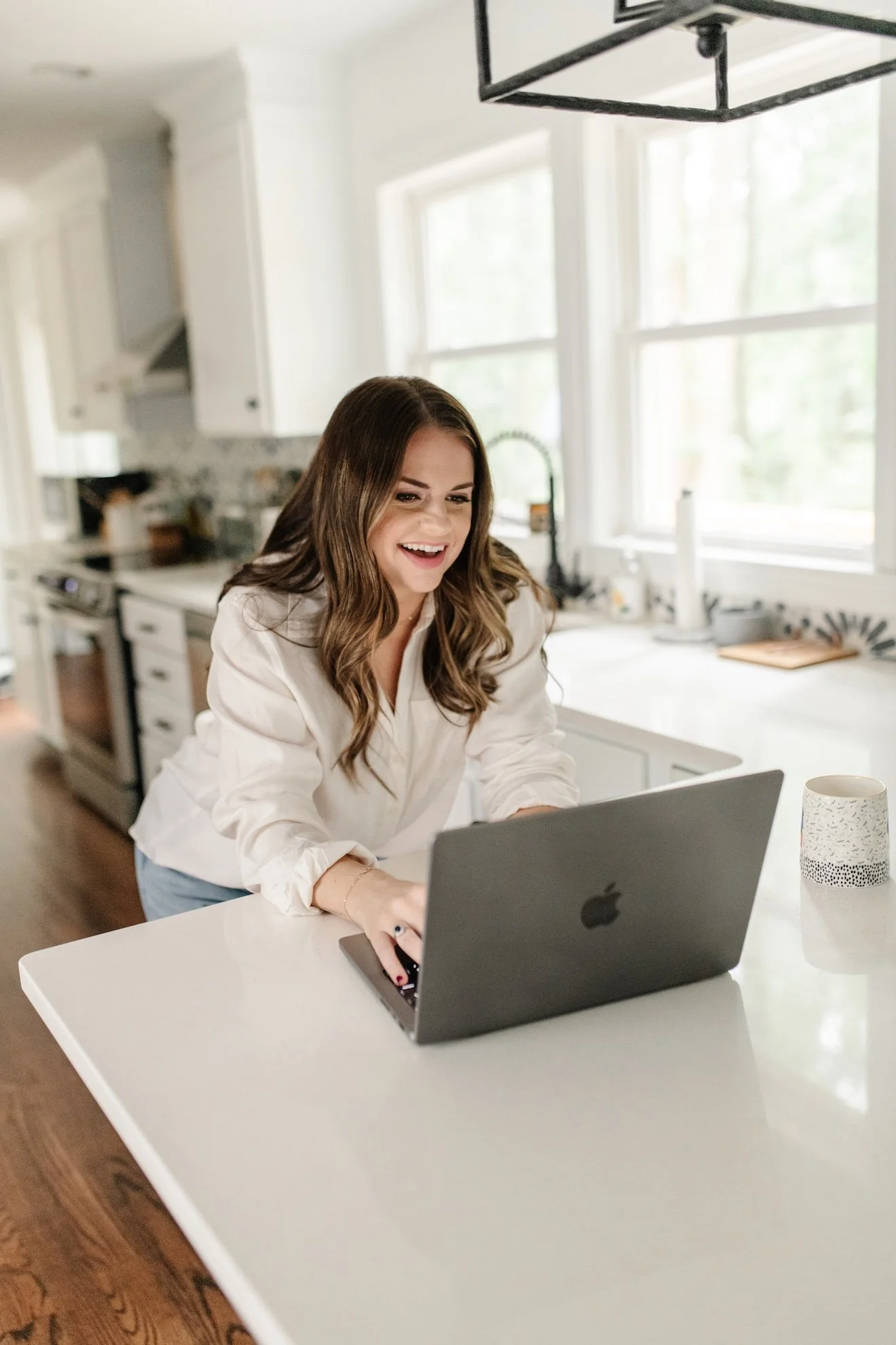 A woman with long brown hair, wearing a white blouse, happily working on a laptop in a bright, modern kitchen.