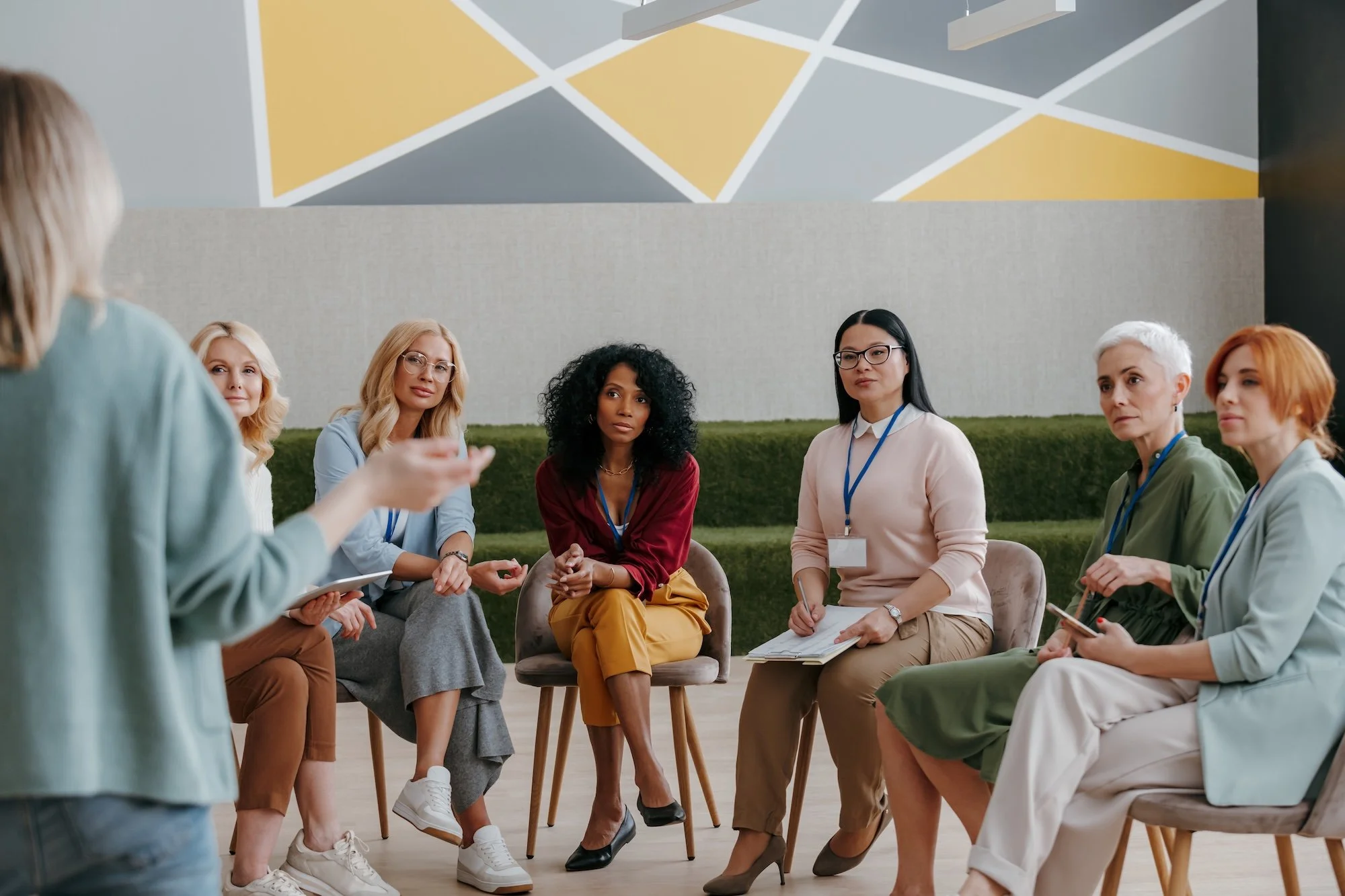 A group of women sitting in a circle during a discussion or meeting inside a room. A woman in the front is standing and speaking, while others listen attentively.