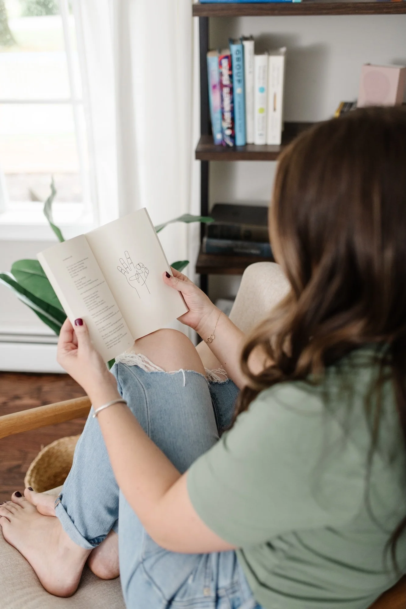 A woman with brown hair reading a book while sitting on a beige couch with her legs up, wearing a green T-shirt and ripped jeans. There is a bookshelf in the background and sunlight coming through a window.