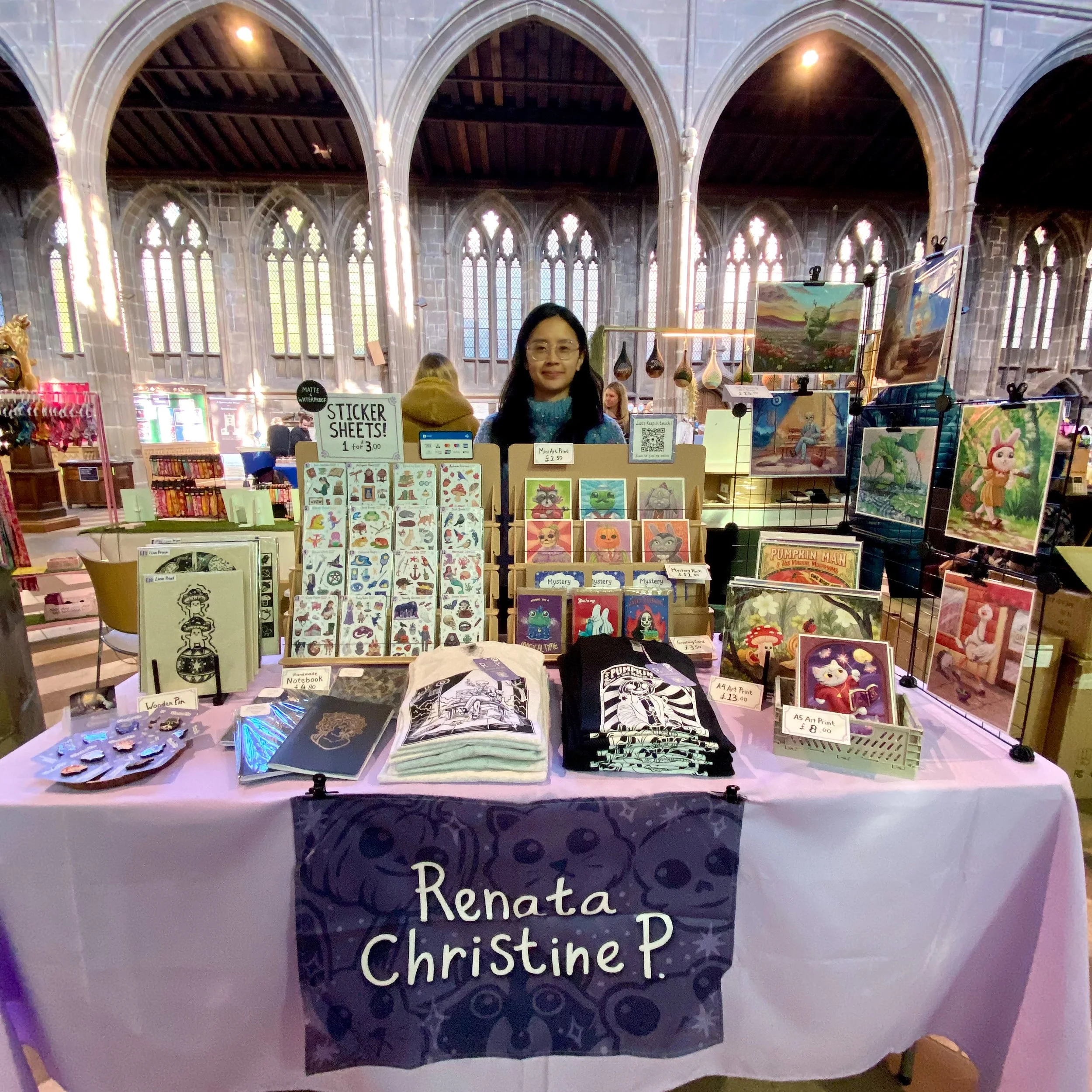 Artist Renata Christine P. at her booth displaying sticker sheets, art prints, notebooks, and tote bags with colorful designs and Halloween themes inside a large, gothic-style building with arched windows.