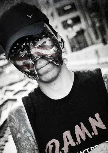 Person wearing a black cap, sunglasses with American flag design, and a bandana covering the lower face, standing outdoors.