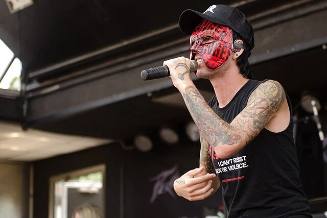A tattooed woman wearing a black cap and a red face mask singing into a microphone on stage.